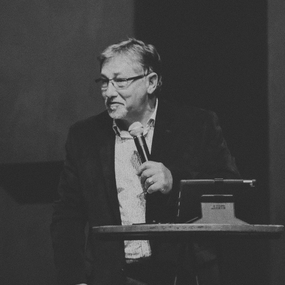 A middle-aged man with glasses and short hair, wearing a suit, holding a microphone and speaking at a podium in a dark room.