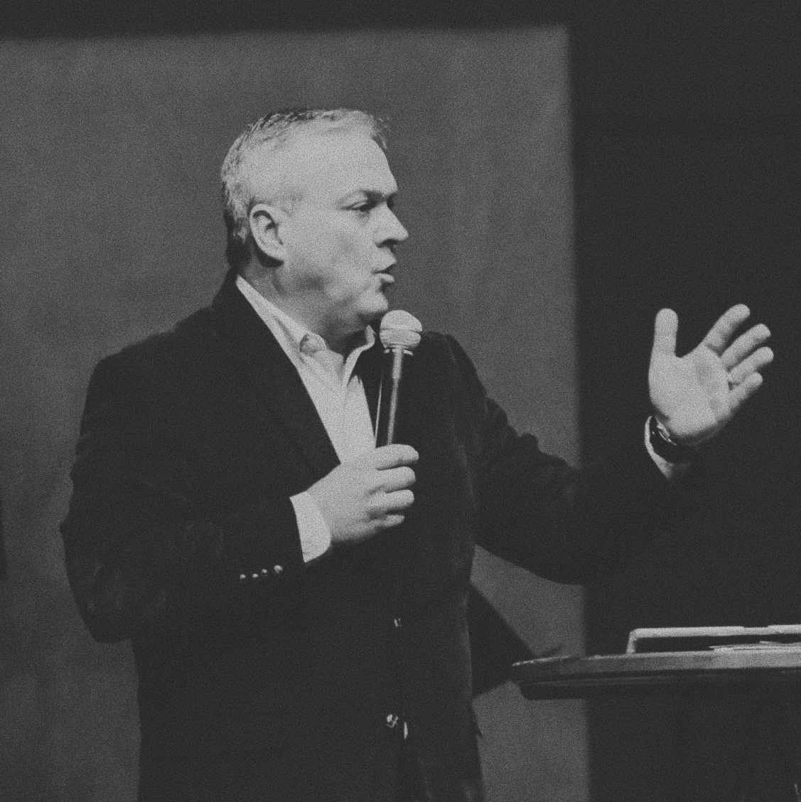 A black-and-white photo of a man in a suit holding a microphone, speaking and gesturing with his right hand, standing behind a podium.