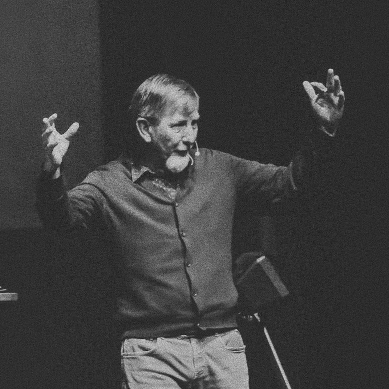 An older man with a beard and mustache, wearing a button-up shirt, is speaking and gesturing with his hands in a dimly lit room.
