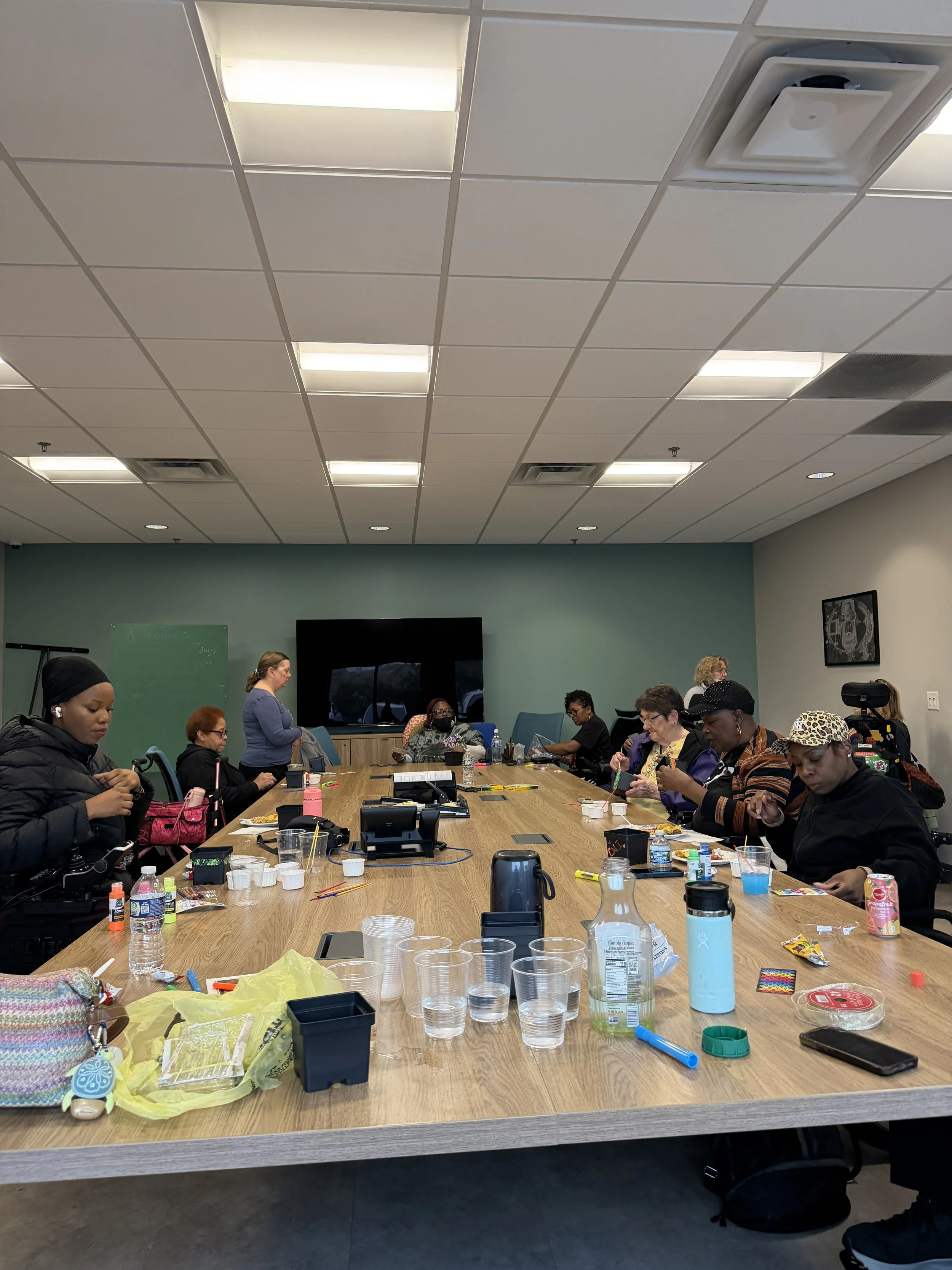 Participants gather around a conference table at Paraquad for a creative group activity, using art supplies and sharing conversation. Several participants use mobility devices.