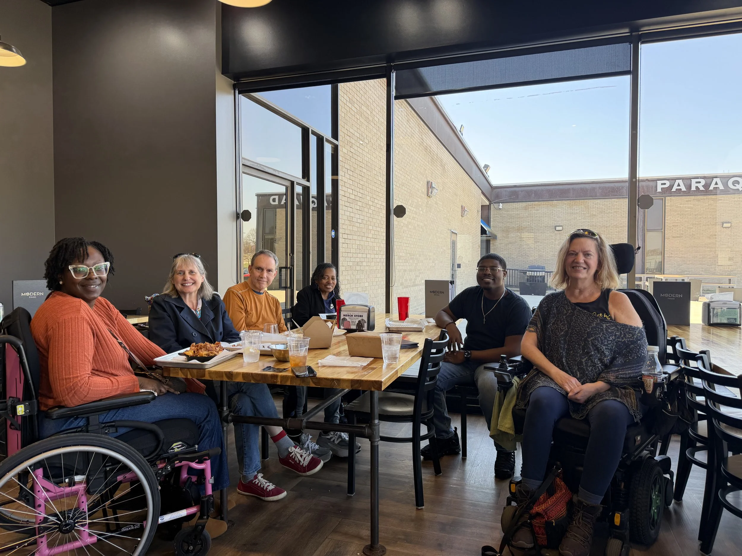Six adults gather around a table at a local brewery during an MS Awareness Month event, sharing a meal and smiling for the camera. Two participants use wheelchairs.