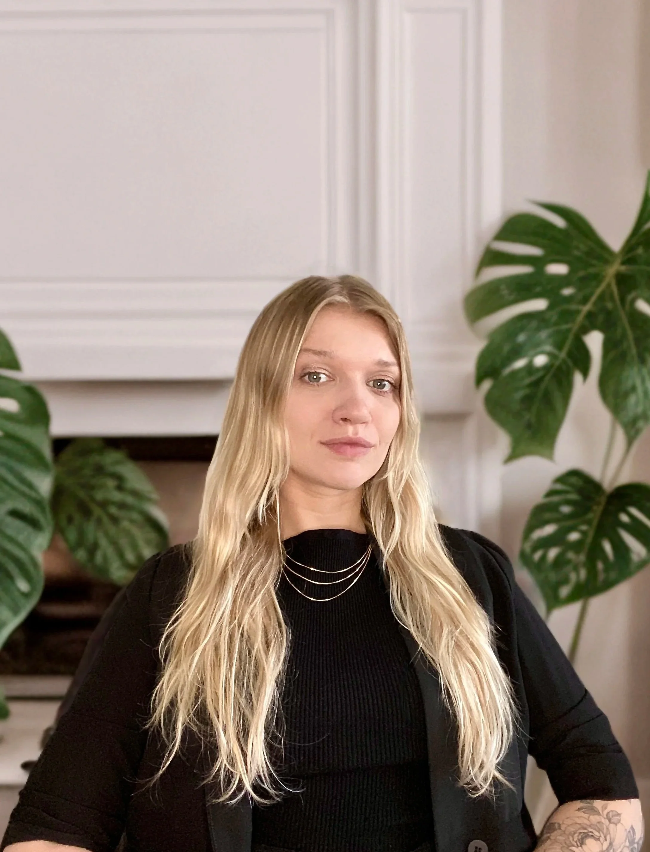 Alex Oumn the Founder of Zen Mondays. A young woman with long blonde hair, wearing a black ribbed top and layered necklaces, sitting in front of white cabinetry and large green houseplants.