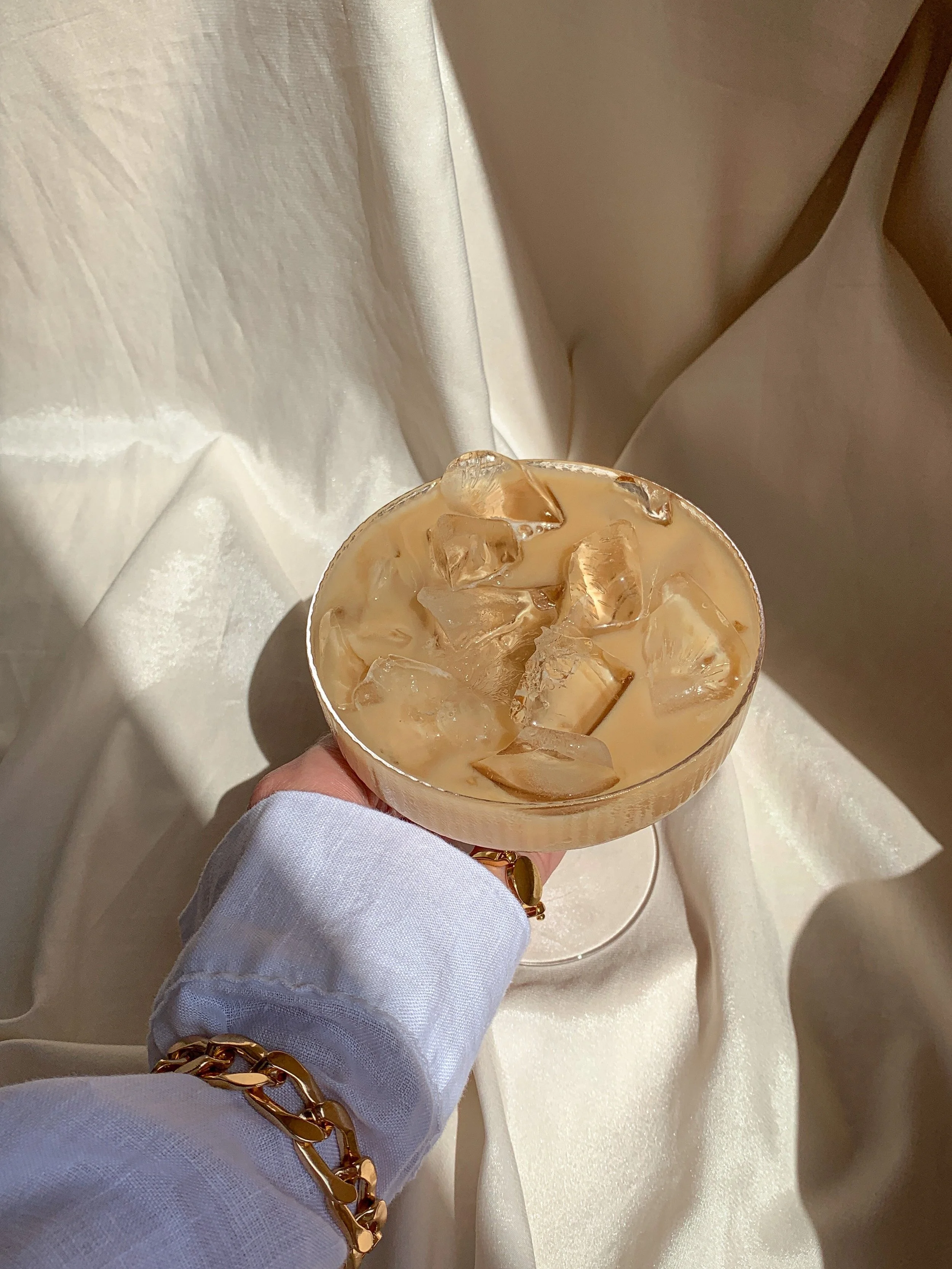 Iced coffee in a glass held by a person wearing a white shirt and gold bracelet, with a draped fabric background.