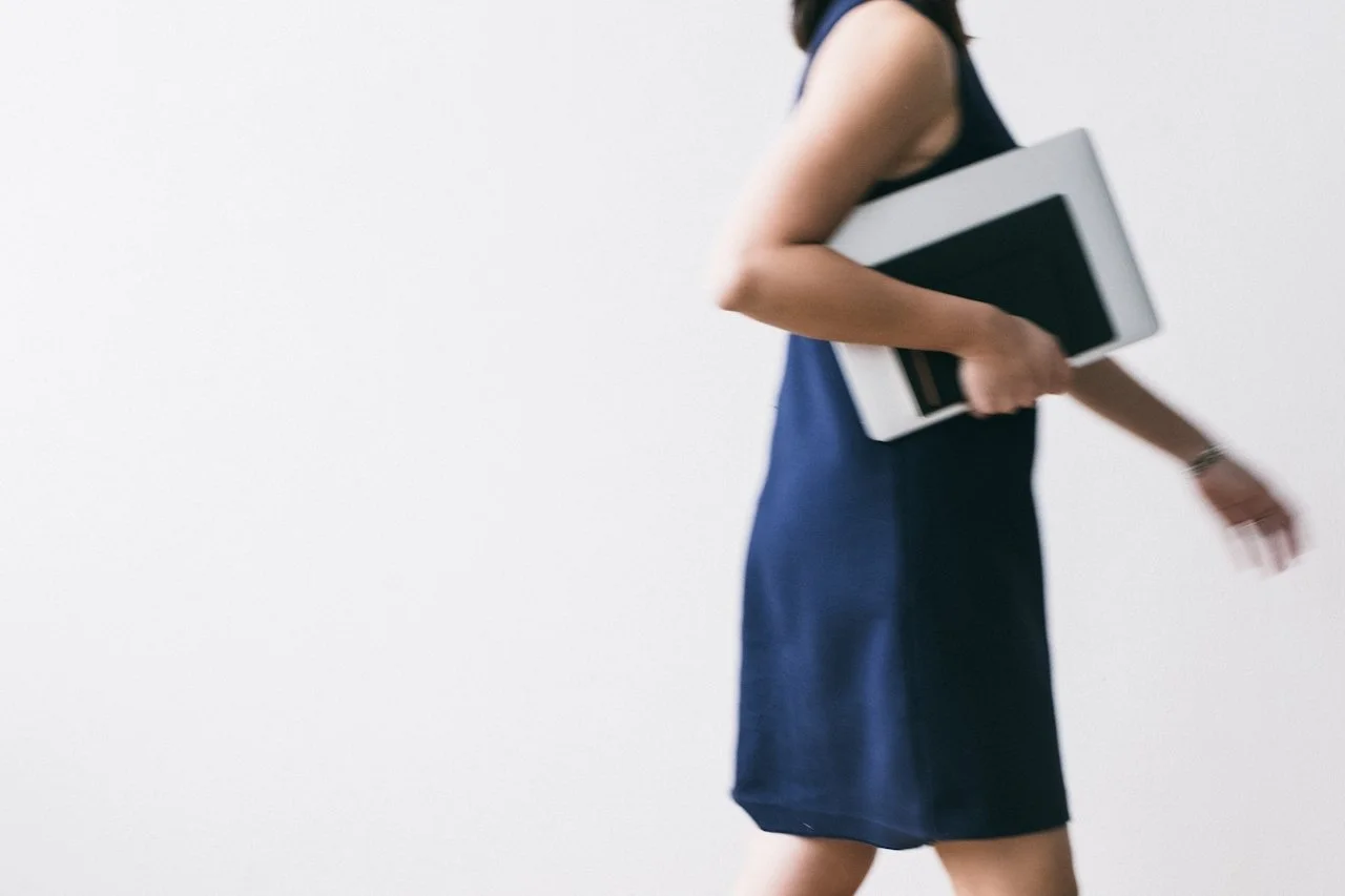 A woman in a blue dress holding a folder or notebook, walking past a plain white wall.