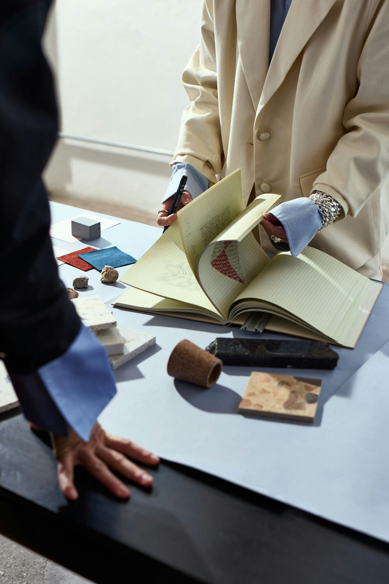 A person in a beige coat and silver jewelry shows a book and various samples on a table during a presentation or discussion.