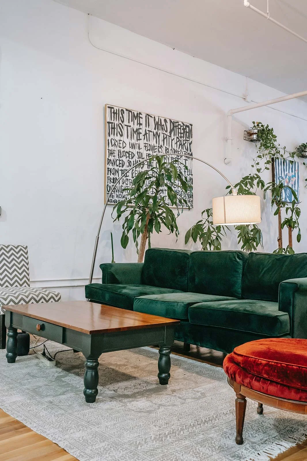 Interior of a living room with a dark green velvet sofa, a wooden coffee table, and a red velvet chair. There are multiple green potted plants and wall art, including a black and white text piece.