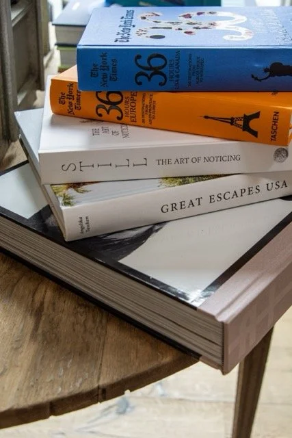 Stack of four travel and photography books on a wooden table, with books titled "The New York Times 36 Hours Europa" in orange, "The New York Times 36 Hours USA & Canada" in blue, "The Art of Noticing" in white, and "Great Escapes USA" in white.
