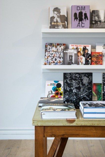 Books and magazines displayed on a small wooden table and white shelves, featuring artwork, photos, and covers with various colors and designs.