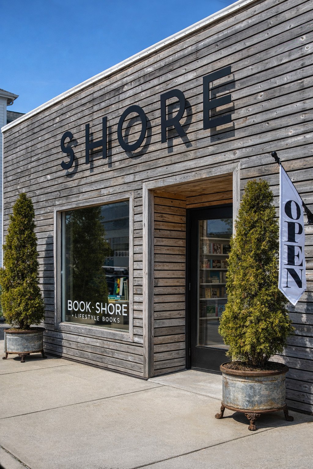 Exterior of a bookstore named Shore with a wooden facade, large black letters spelling Shore, a window with books inside, and potted trees outside. A sign next to the door reads Open.