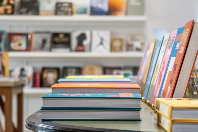 Stack of colorful books on a table with a bookshelf in the background.