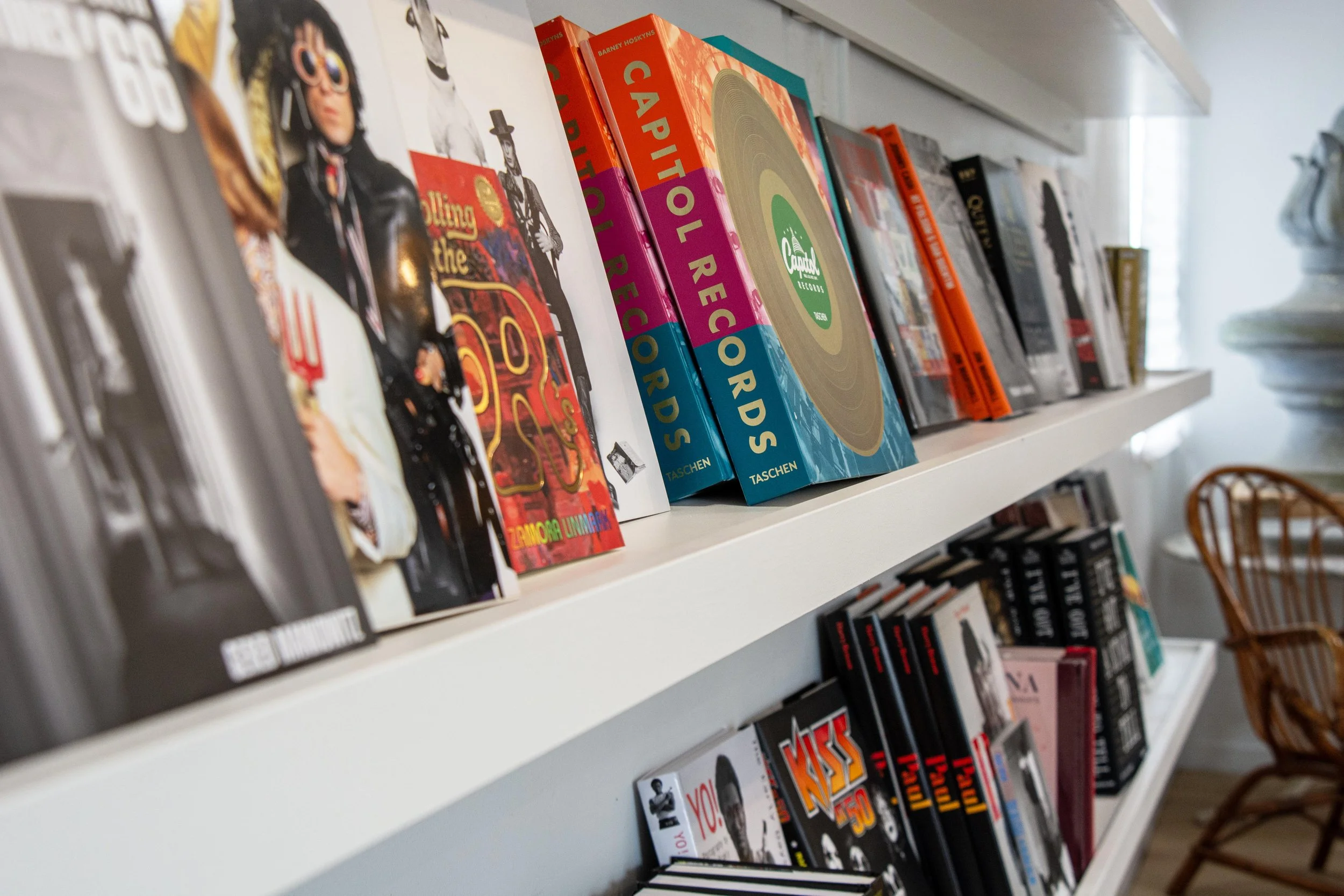 Books and magazines on white shelves in a room, including titles like 'Capitol Records' and 'KST 50' with a wicker chair nearby.