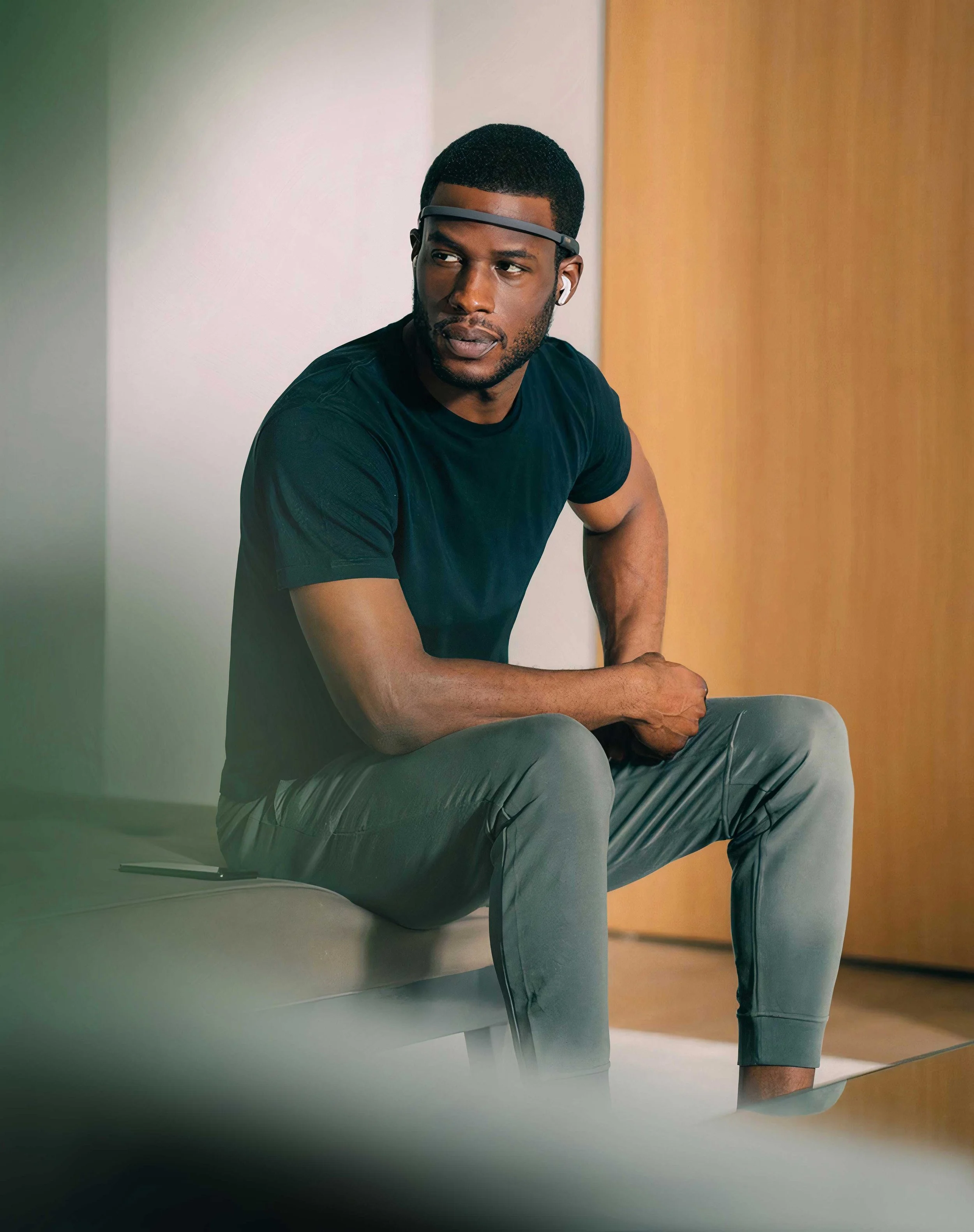 A man wearing a black t-shirt, gray pants, and wireless earbuds, sitting on a gray chair indoors, looking to the side.