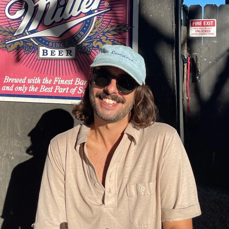 Man with long hair and beard wearing sunglasses, a light beige shirt, and a light blue baseball cap, standing outside near a dark wall with a sign and a fire exit door.