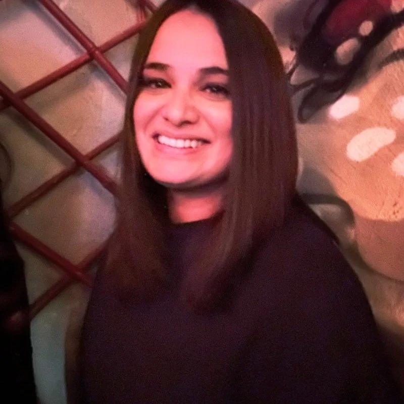 A woman with shoulder-length dark hair smiling, wearing a black top, sitting in a cozy, warmly lit setting with a lattice-like wooden divider and a patterned carpet in the background.