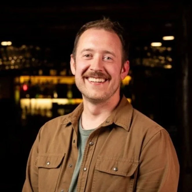 A smiling man with a mustache and short hair, wearing a brown jacket and a gray shirt, standing outdoors at night with city lights in the background.