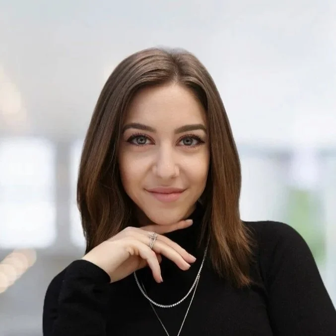 A young woman with shoulder-length brown hair, wearing a black top and silver jewelry, smiling and resting her chin on her hand in an indoor setting with blurred windows in the background.