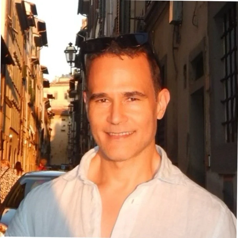 A man smiling outdoors on a narrow city street with buildings on both sides and a vintage-style lamp post in the background.