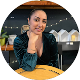 A woman with dark hair styled in a bun sitting at a table in a modern indoor space, smiling at the camera.