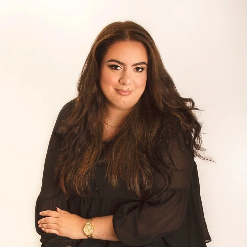 A woman with long brown wavy hair wearing a black top with sheer sleeves, a gold watch, and a delicate necklace, standing against a plain white background.