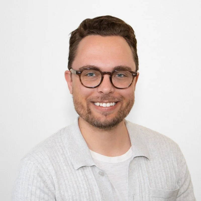 Headshot of a smiling man with glasses, light-colored shirt, and a white background.