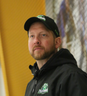 A man with a beard wearing a black jacket and cap, standing near a chain-link fence.