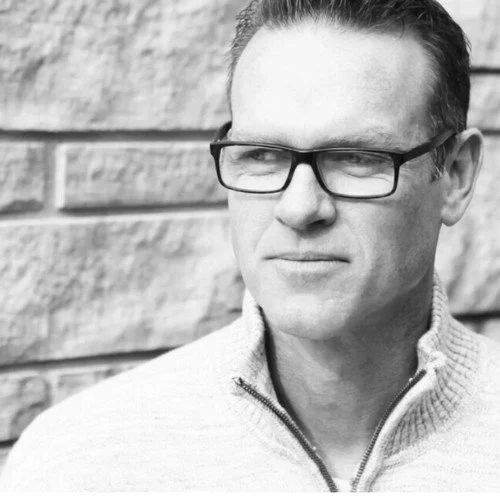 Black and white photo of a man wearing glasses and a zip-up sweater, standing against a brick wall, looking to the side.
