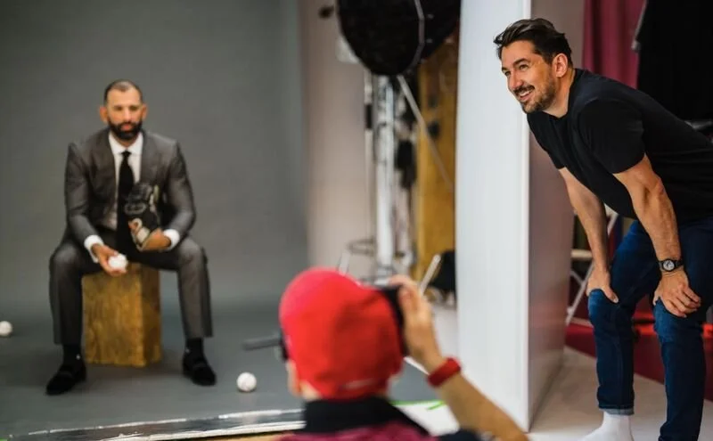 Photo of a photoshoot in a studio with a man in a suit sitting on a wooden box and a man in casual clothes leaning from behind a white wall, both smiling, with a photographer in a red hat and casual outfit in the foreground.