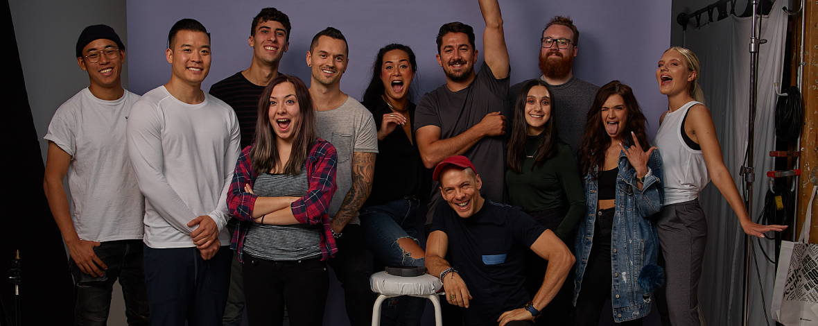 Group of 11 young adults posing in a photo studio, smiling and making playful gestures.