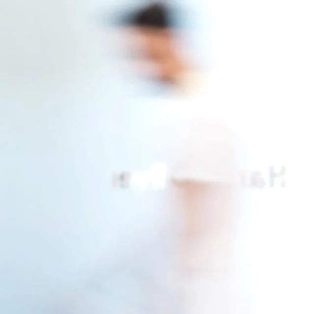 A blurred person with dark hair wearing a white shirt standing in front of a light background.