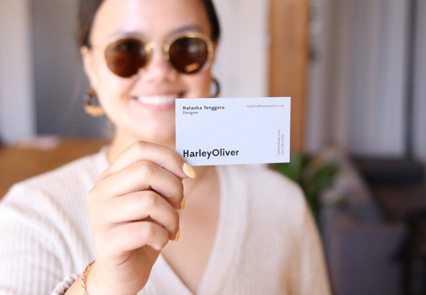 Woman wearing sunglasses holding a business card with the text 'HarleyOliver' in focus, smiling