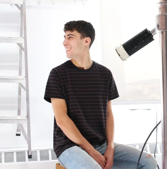 Young man smiling and sitting on a stool in a studio under a photography spotlight with a ladder nearby.