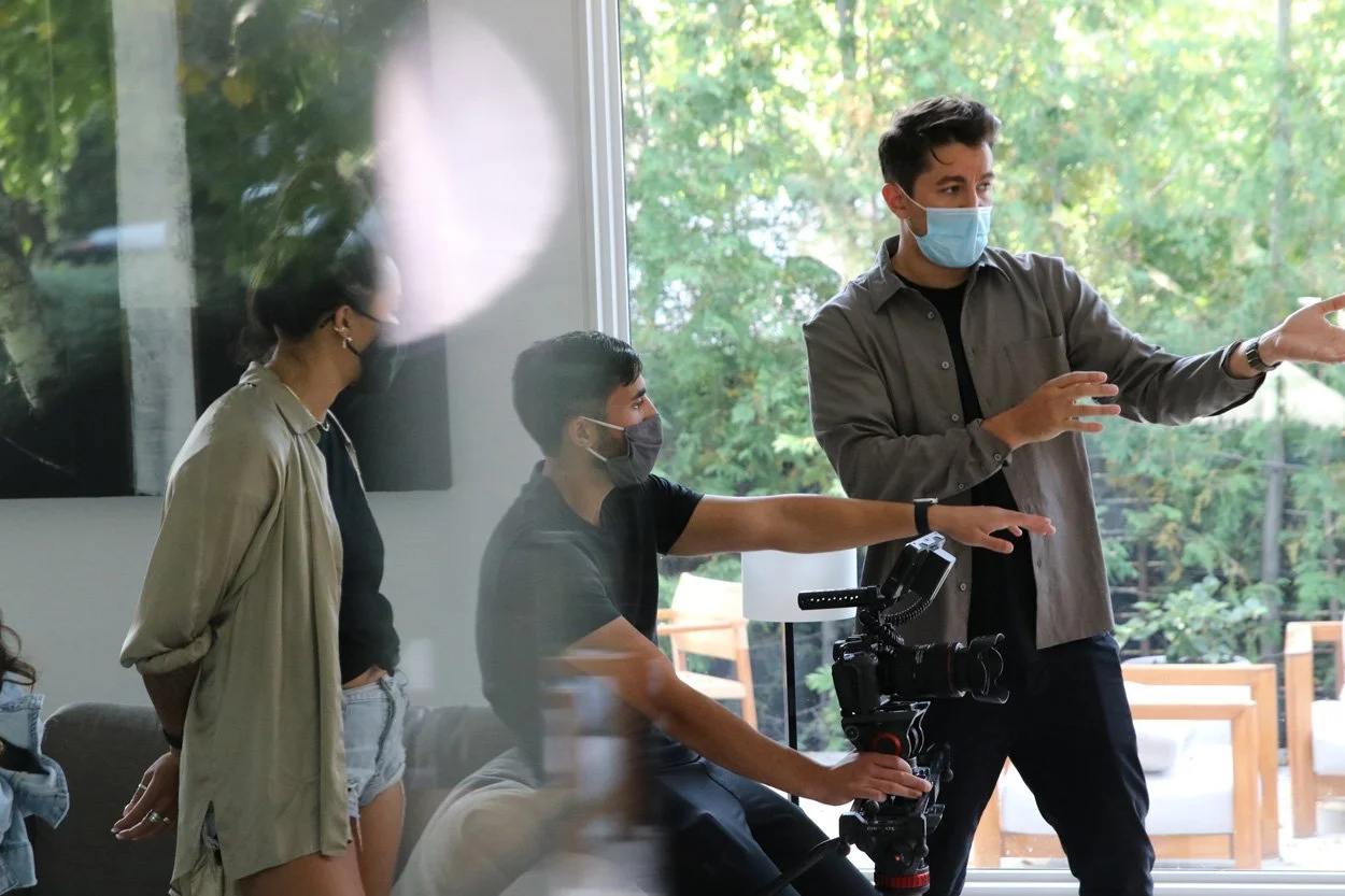 Three people wearing masks in a room with large windows, two are standing, one seated with a camera, engaged in a discussion or filming.
