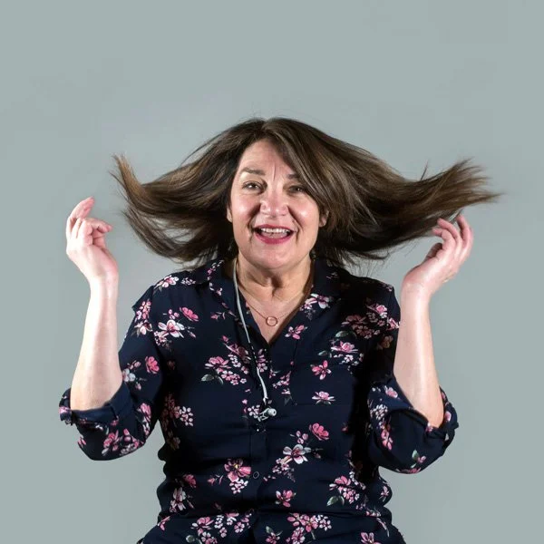 Mid-aged woman with long brown hair, wearing a floral navy blouse, smiling, with hair flying around her head against a plain gray background.