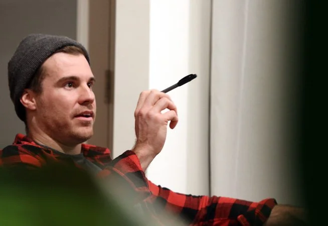 A man wearing a gray beanie and red plaid shirt holding a black marker or pen, sitting and looking at a whiteboard or screen in a room.