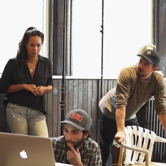 Three people in a room with desk and laptop, two men and one woman. The woman stands and looks at the computer, while the men sit and look at the screen.