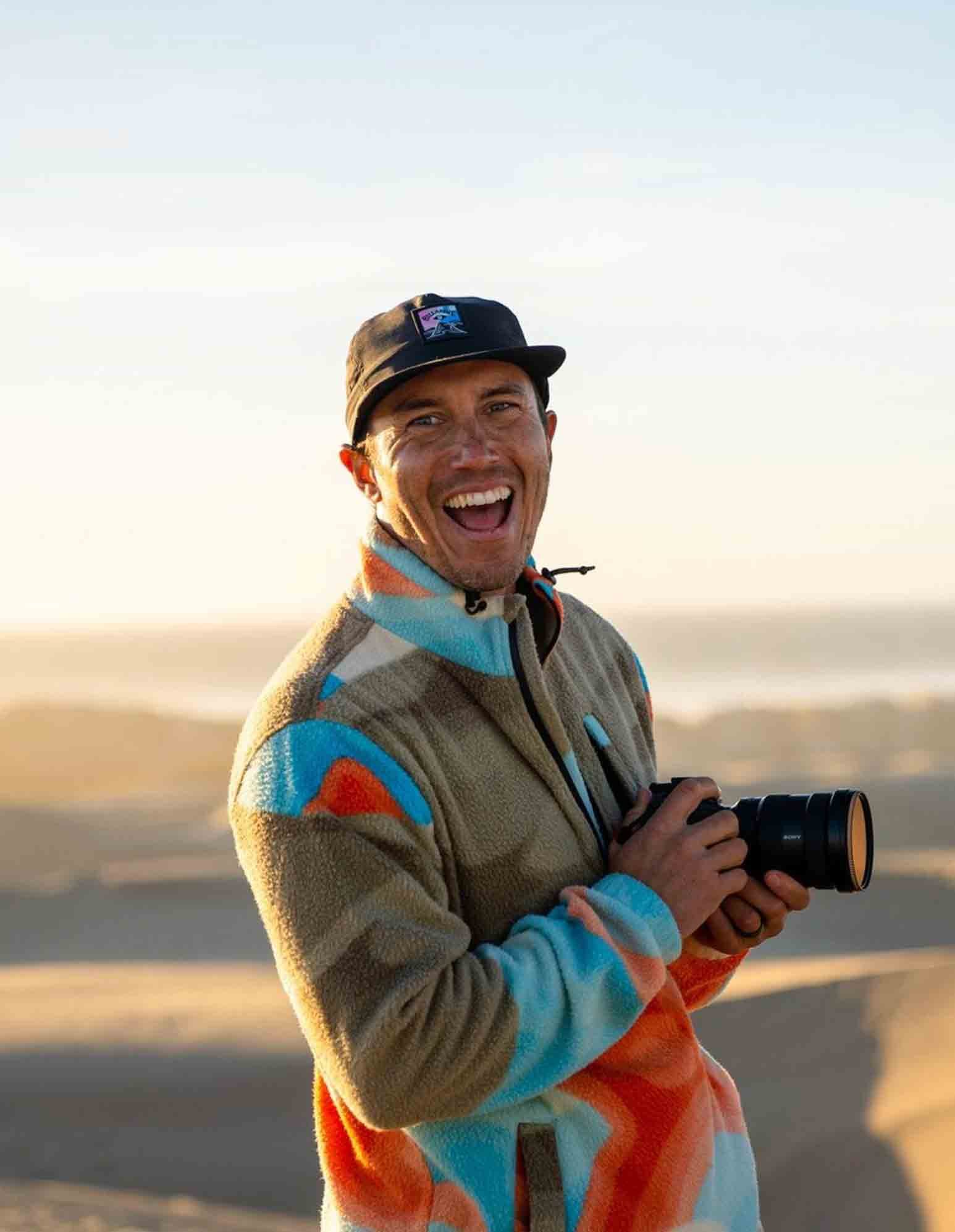 A man in colorful outdoor clothing smiling and holding a camera at the beach during sunset.