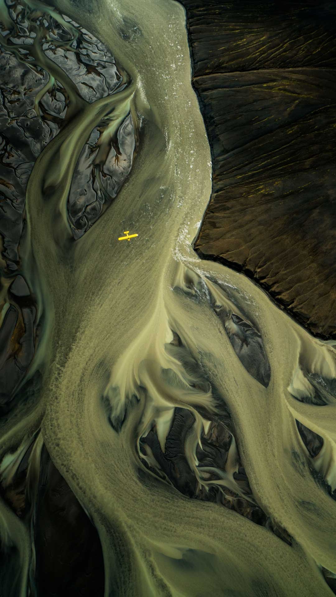 Aerial view of a winding river with black and greenish water, flows through a rugged landscape with steep cliffs and dark terrain.