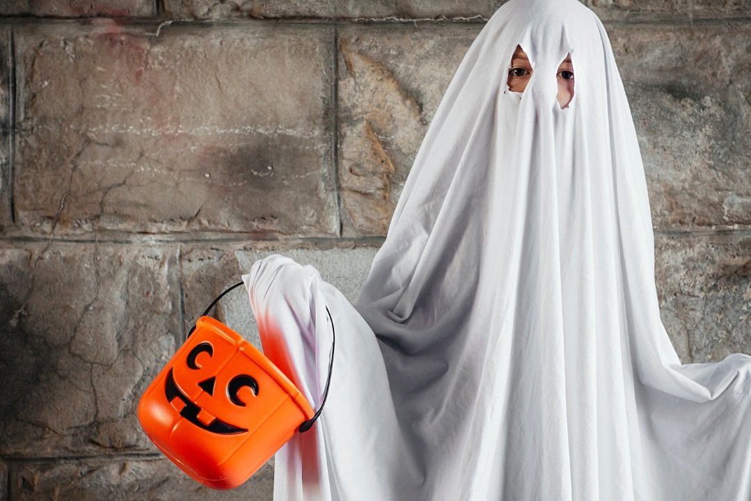 Child dressed as a ghost for Halloween, holding an orange pumpkin-shaped bucket with a smiling jack-o'-lantern face, in front of a stone wall.