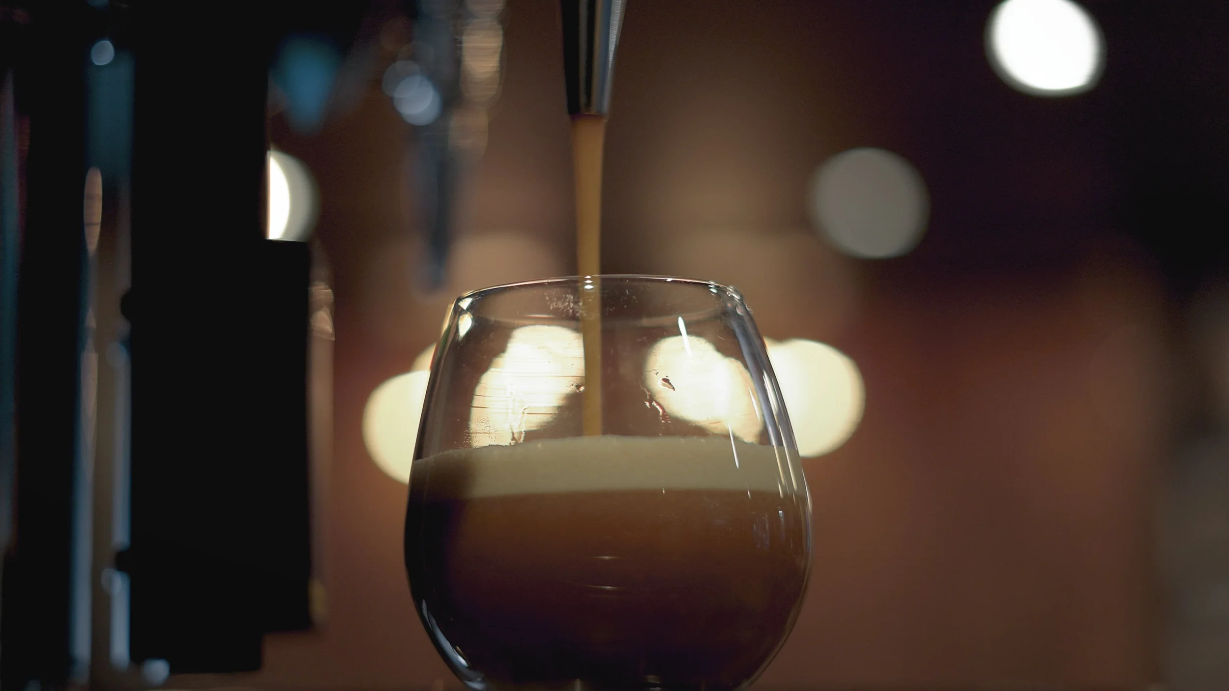 A tap pouring a dark beer into a rounded glass, with a frothy head forming at the top, against a blurred background of warm lights.