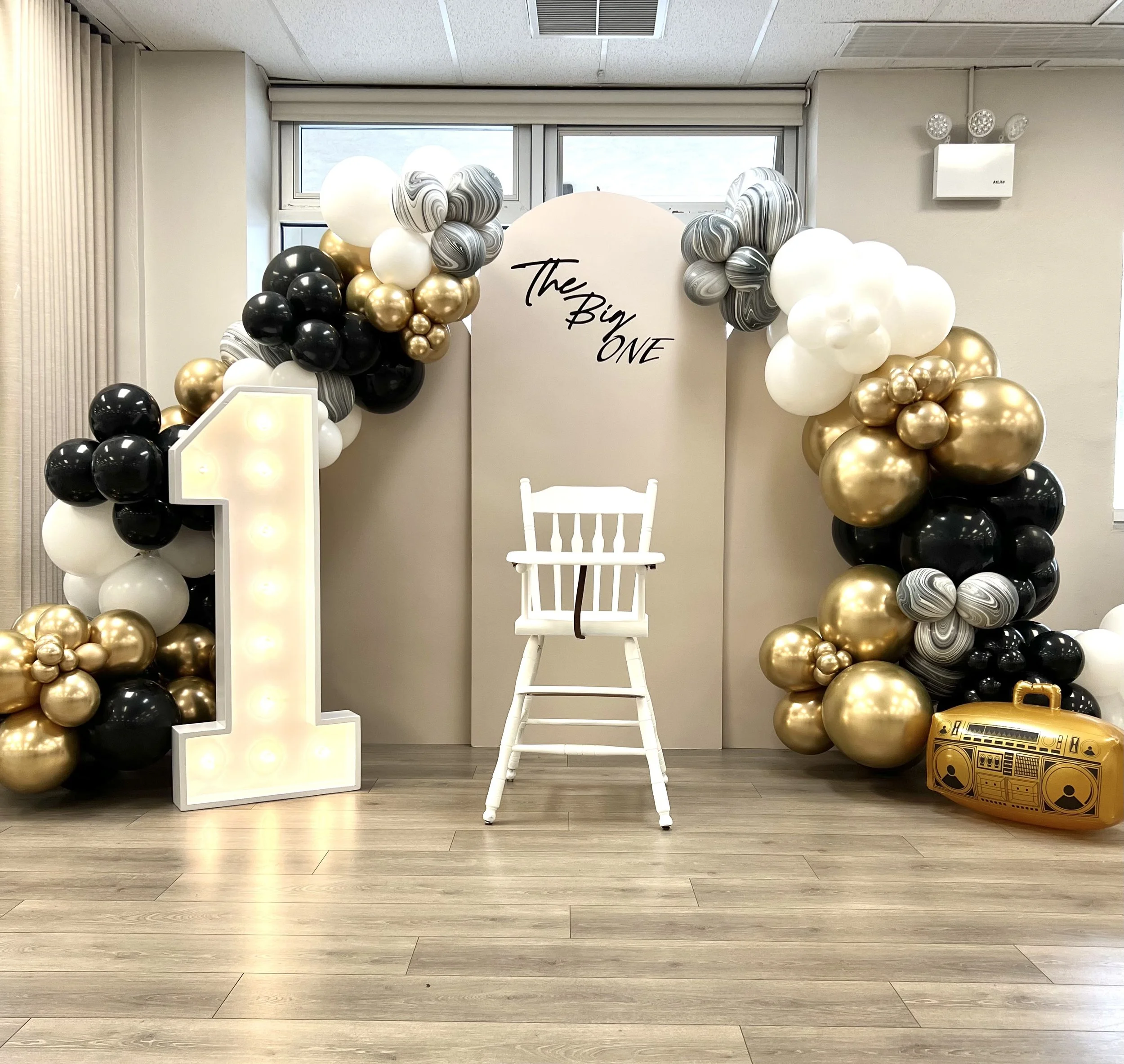 Decorations for a first birthday celebration, featuring a large illuminated number one, a white high chair, a backdrop with the words 'The Big ONE,' and a balloon arch with black, white, gold, and marbled balloons.