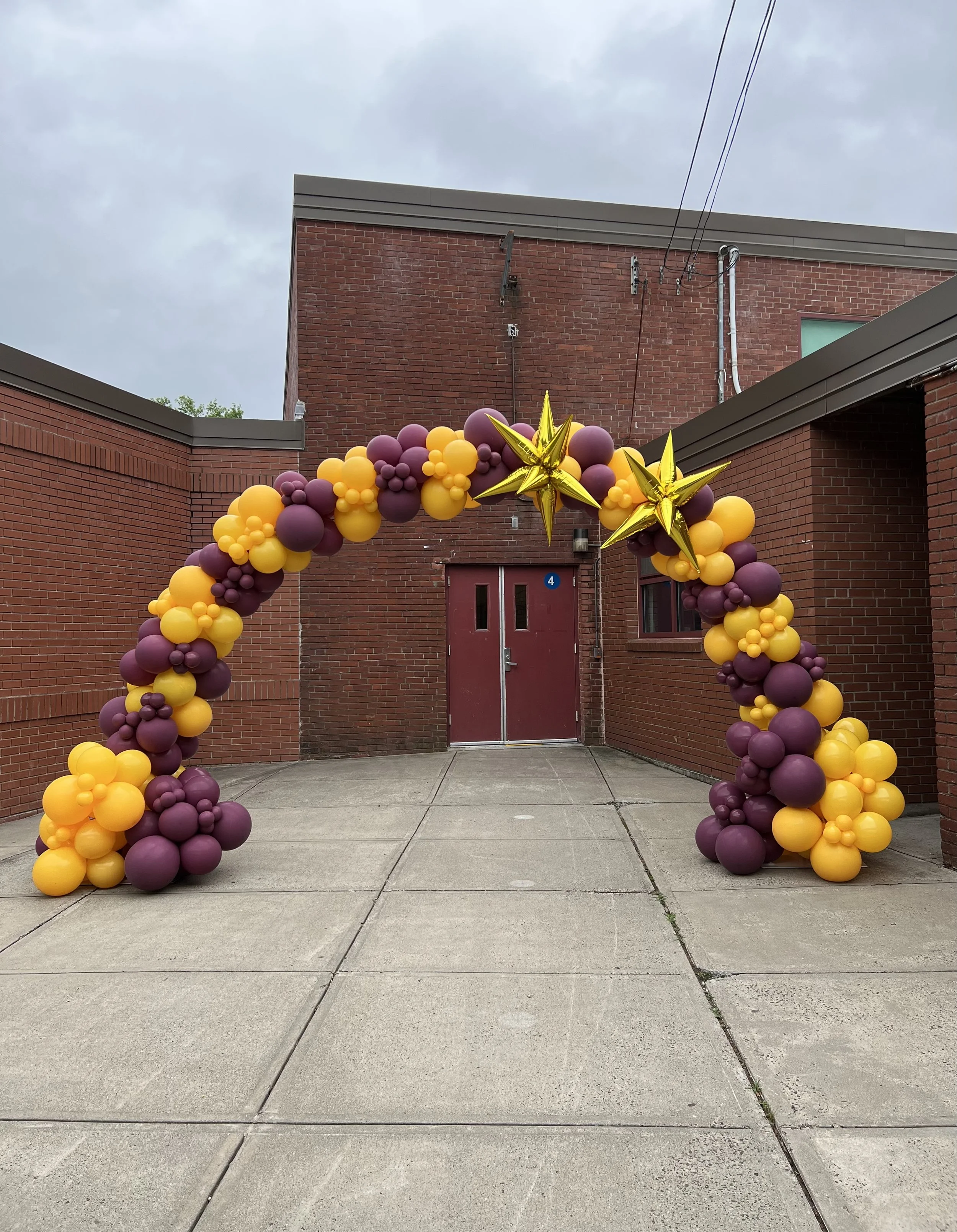 Balloon arch made with purple and yellow balloons, decorated with large gold star-shaped balloons, set up outside a brick building with a red door, under an overcast sky.
