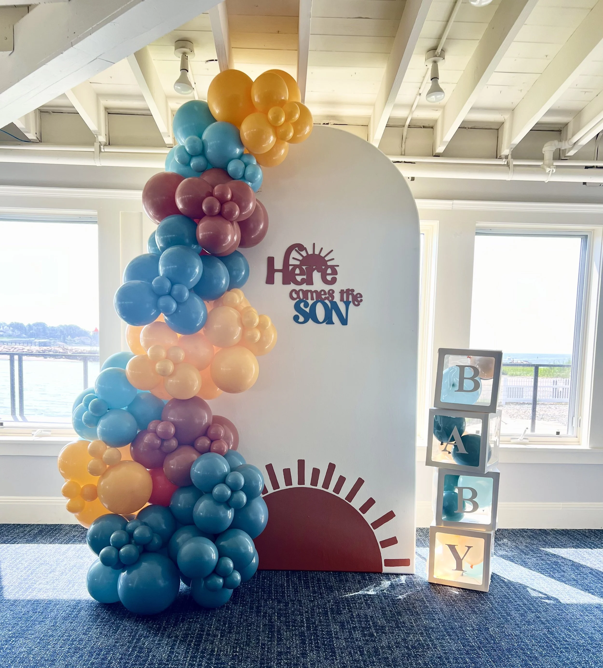 Decorative display for a baby shower with colorful balloons arranged on the left side and a white standing board with the phrase 'Here comes the SON' in the center. To the right, there are four stacked cubes with the letters B, A, B, and Y, illuminat