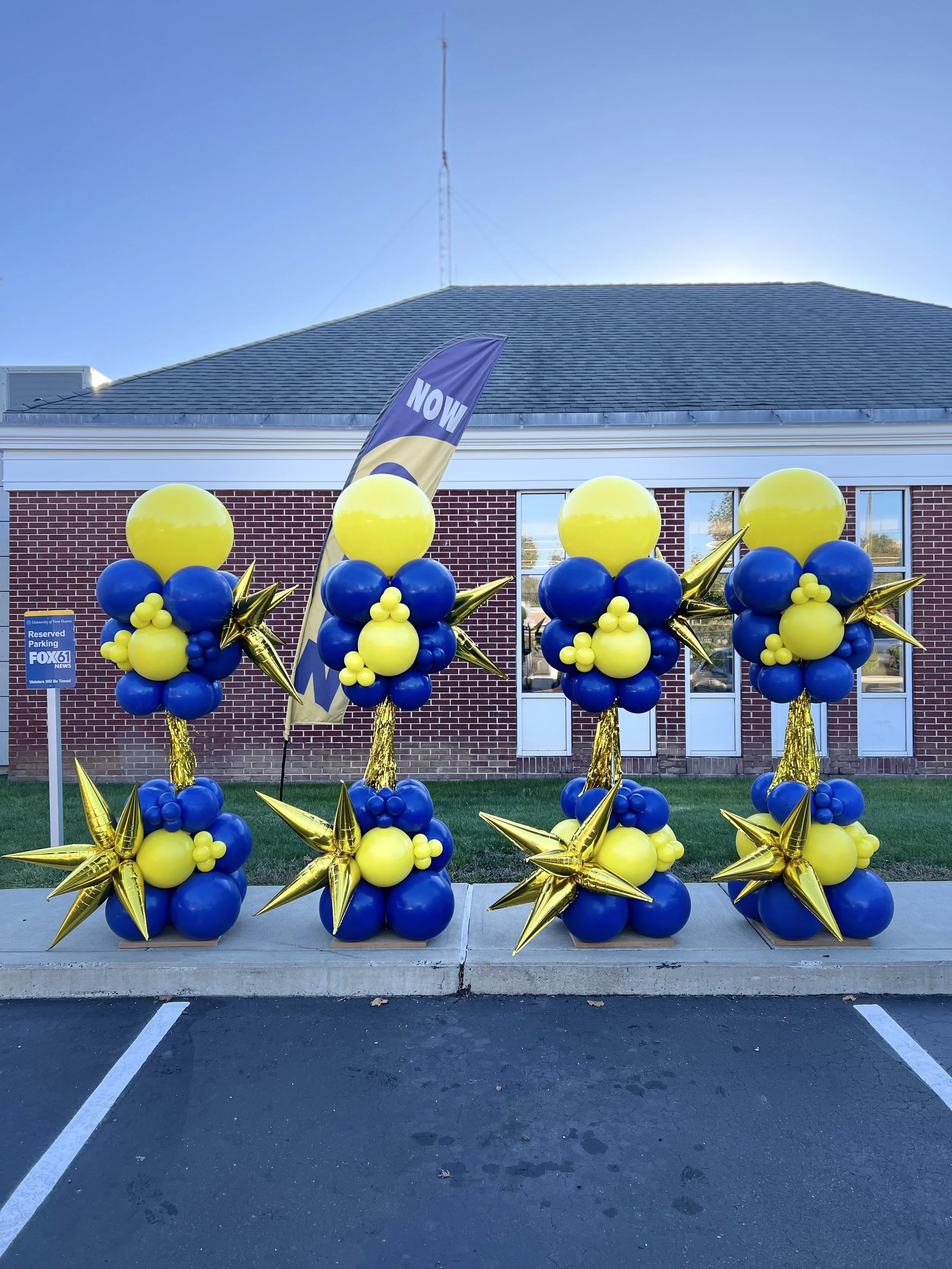 Four balloon arrangements with yellow and blue balloons, gold stars, and a black flag with yellow accents that reads "NOW" outside a brick building with a parking lot.