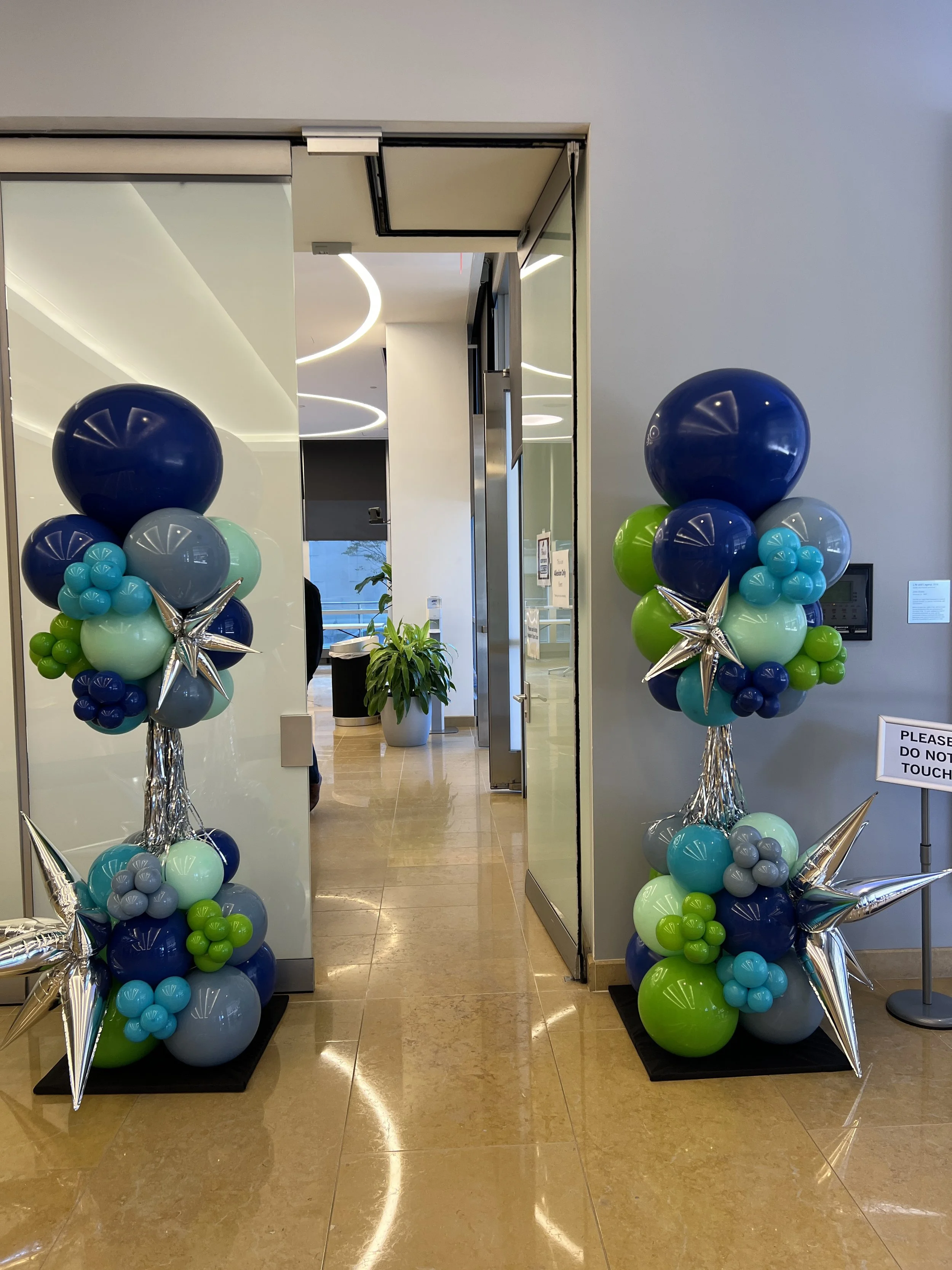 Two balloon arrangements with large blue, green, and gray balloons, accented with silver star-shaped balloons, placed on black bases inside a building entrance.