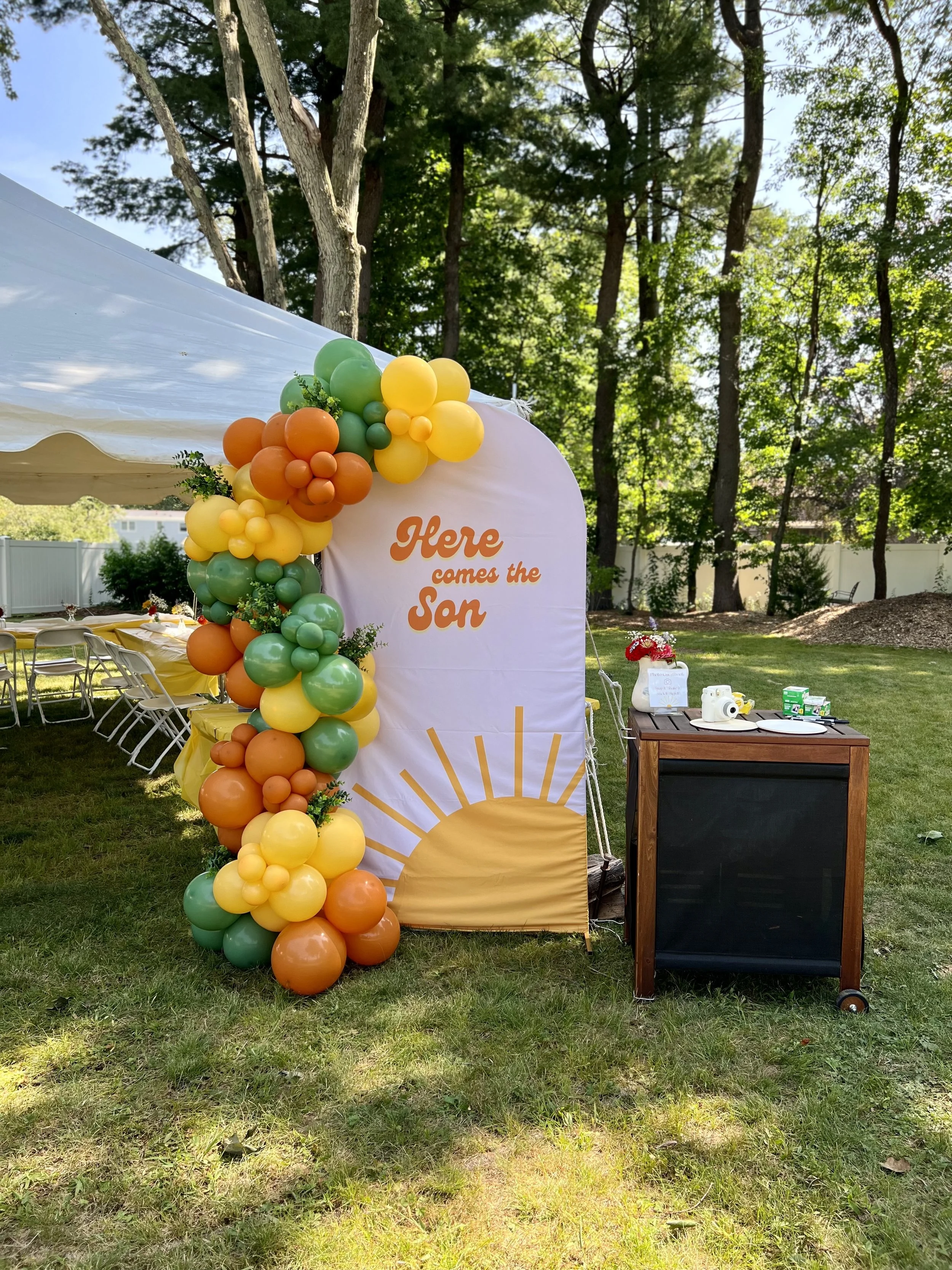 Decorative outdoor setup with a balloon arch in orange, yellow, and green, a white backdrop with the words "Here comes the Son" and a sunrise graphic, and a small wooden table with camera props, on a grassy area surrounded by trees.
