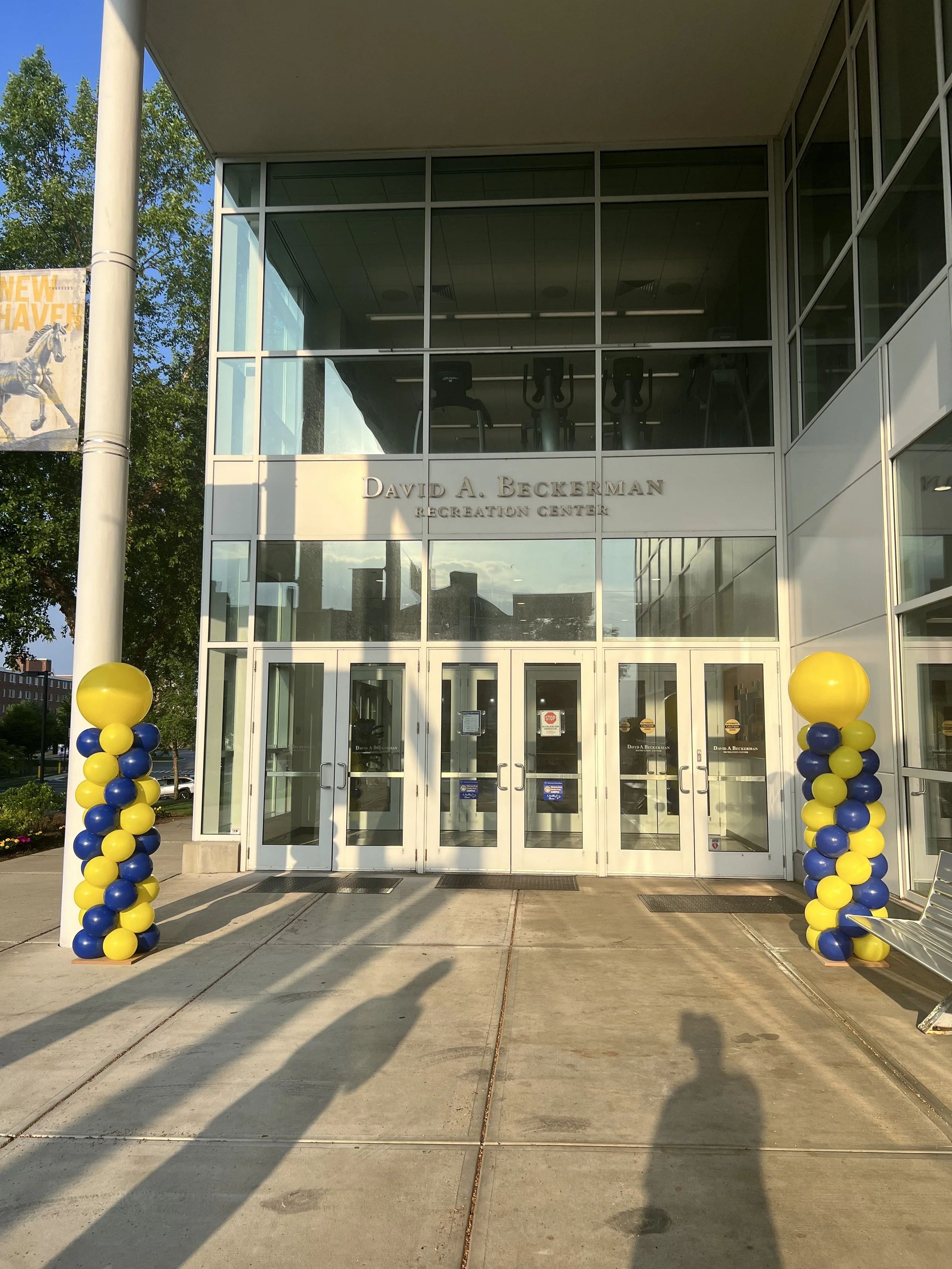 Entrance of the David A. Beckerman Recreation Center decorated with yellow and blue balloons on either side of the doors, with glass doors and windows showing gym equipment inside.