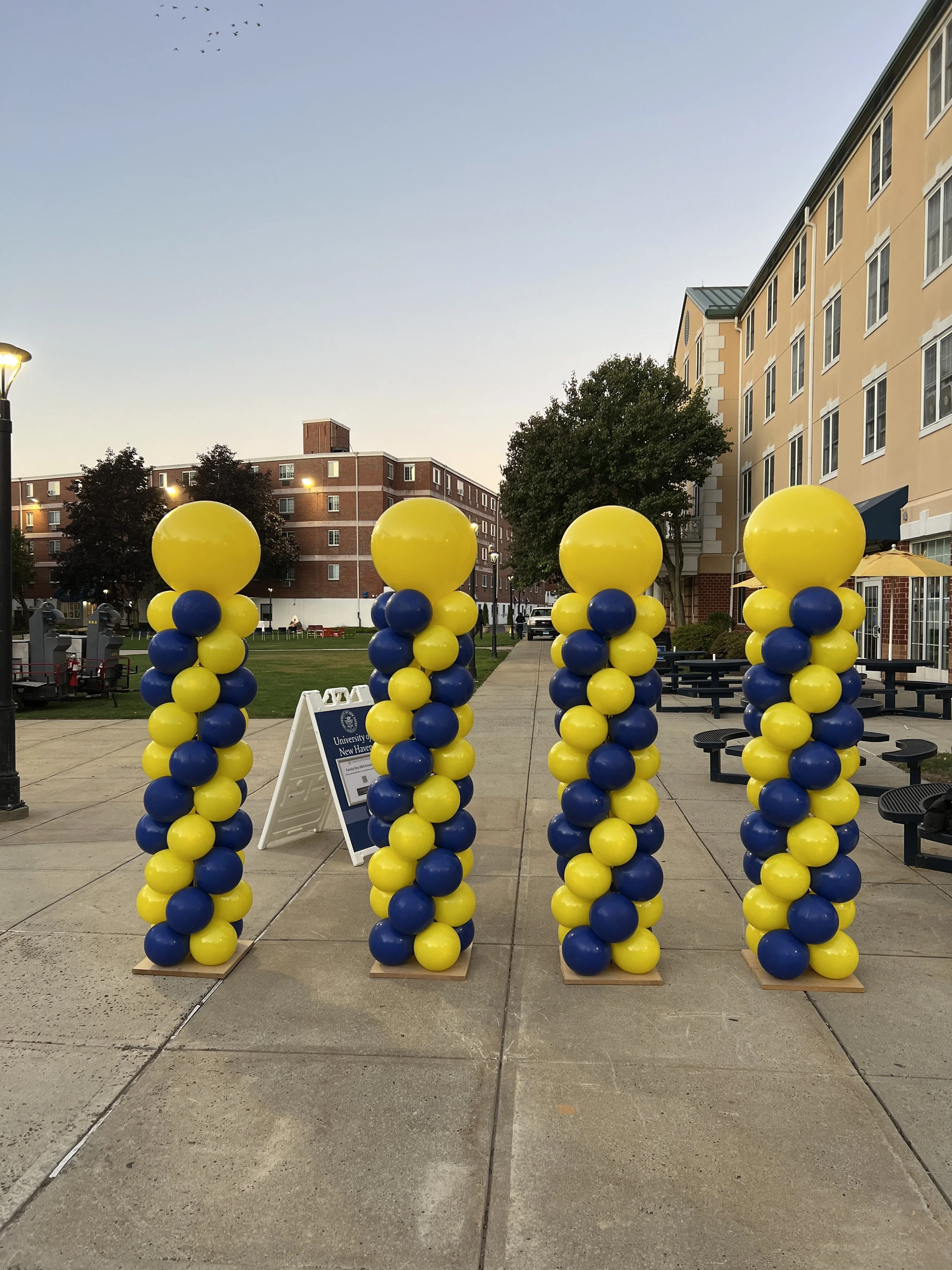 Four columns of yellow and blue balloons placed along a sidewalk outside a campus or residential building, with a signboard in front of them.