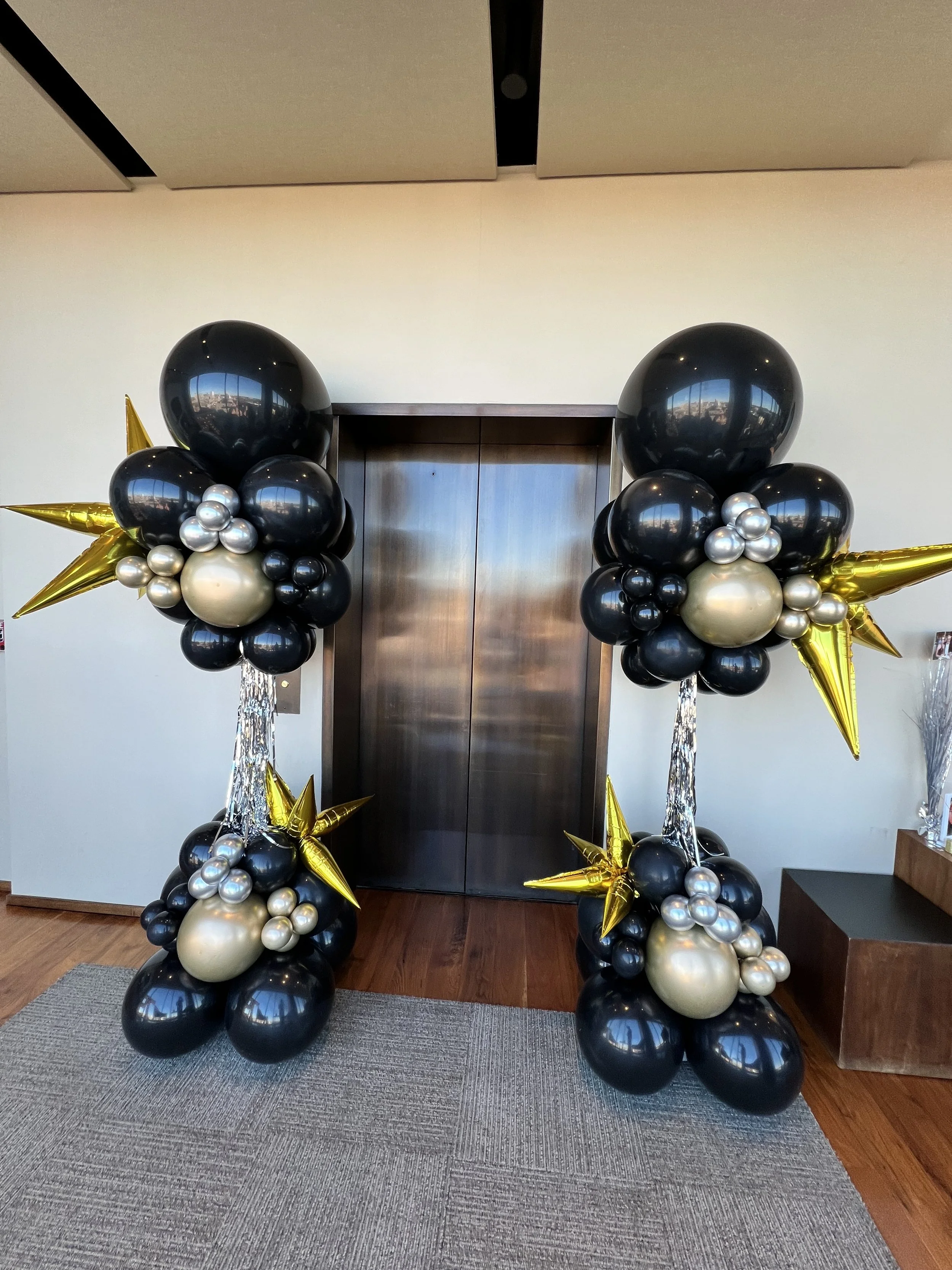 Balloon arch with black, gold, and silver balloons, featuring star-shaped balloons and reflective surfaces, positioned in front of an elevator door.