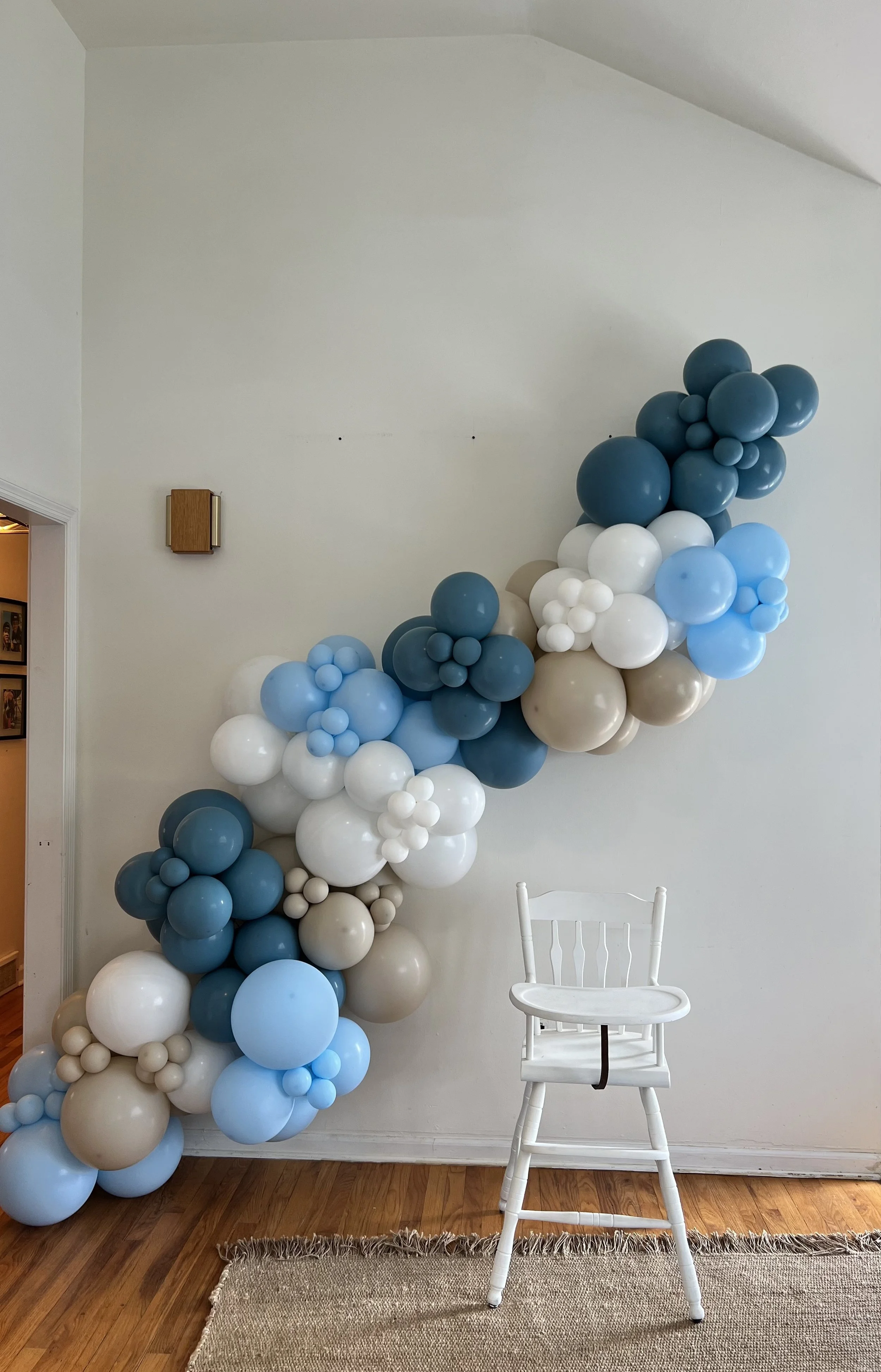 Decorative balloon arrangement in shades of blue, white, and beige arranged in a cascading fashion against a white wall, with a white wooden chair in front of it and a beige rug on a wooden floor.