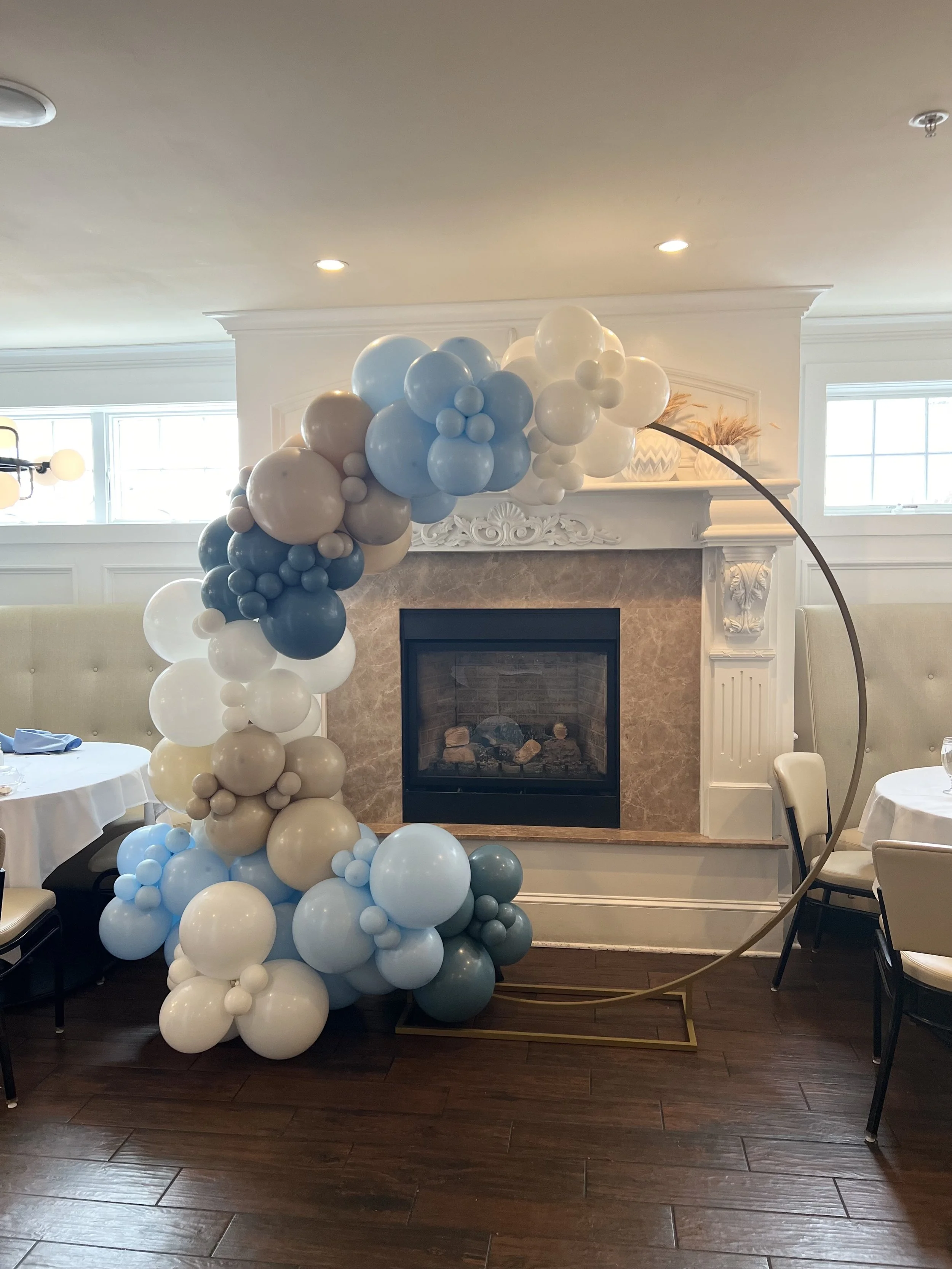 Decorative balloon arch in shades of blue, white, cream, and beige in front of a fireplace in a dining area with round tables and chairs.